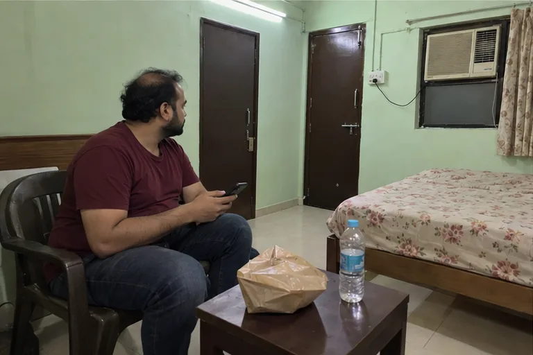 A man in a maroon T-shirt waits on a plastic chair in a cheap lodge room late at night, phone in hand