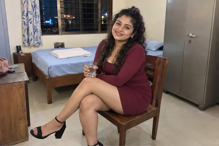 A woman in a maroon short dress sits on a chair in a clean one-room Kurla flat, a glass of water in her hand